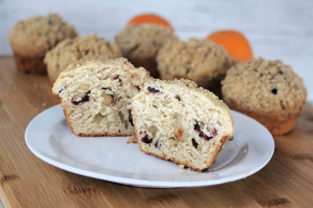 A dried cranberry muffins cut in half on a white plate with more muffins in the background with oranges.