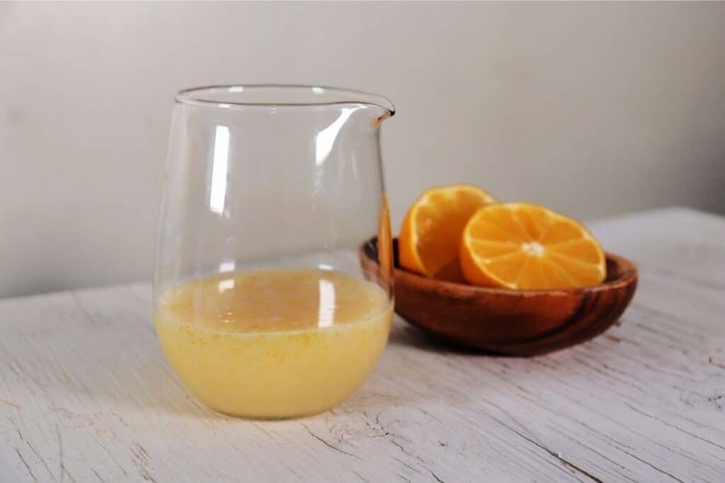 Lemon glaze in a clear glass pitcher in front of a wooden bowl with cut open lemons.