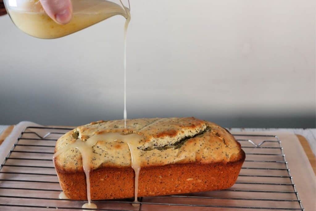 Lemon glaze being poured out of a glass pitcher onto a loaf of lemon poppyseed bread sitting on a wire rack.
