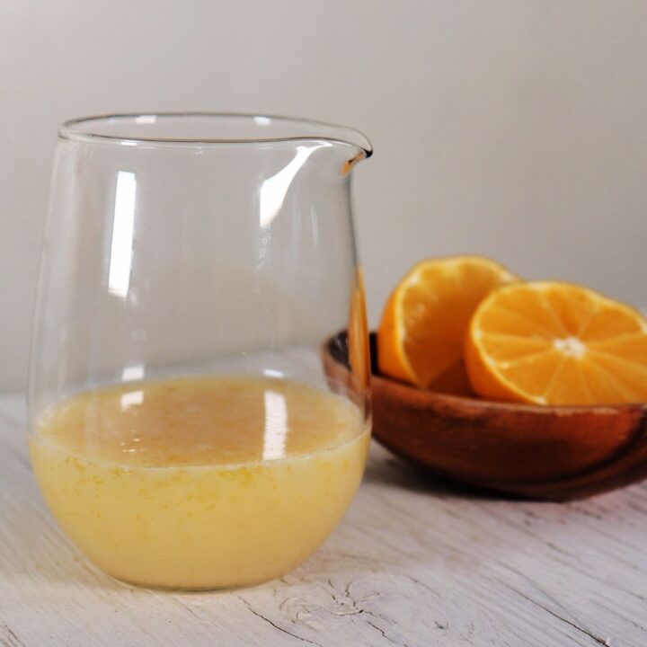 Lemon glaze in a clear glass pitcher in front of a wooden bowl with cut open lemons.