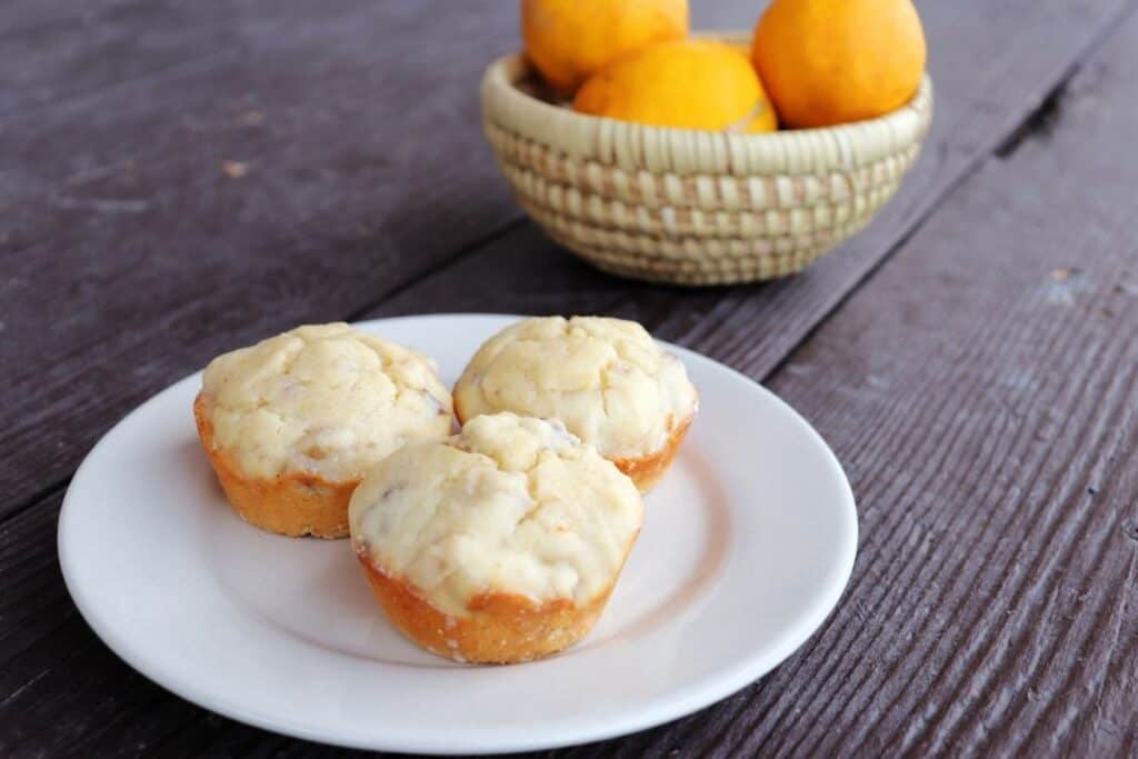 3 lemon muffins on a white plate with a basket fresh meyer lemons in the background.