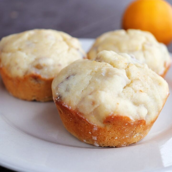 3 lemon muffins on a white plate with a fresh meyer lemon in the background.