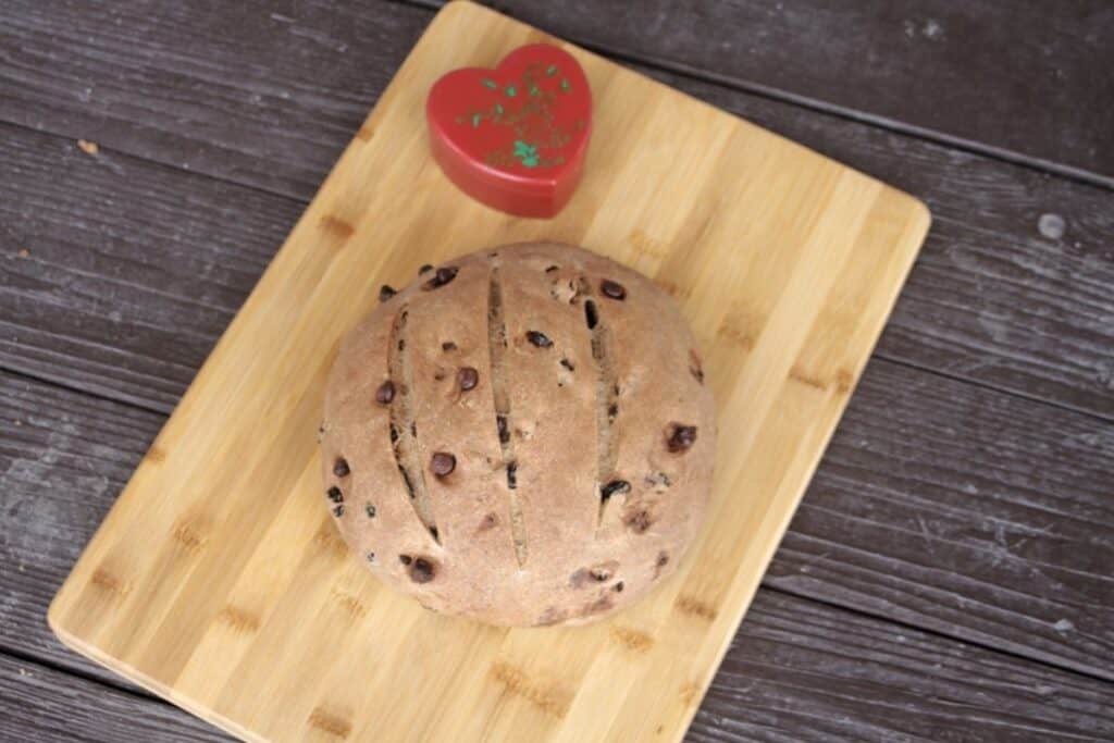 A round loaf of pumpernickel raisin bread on a wood cutting board with a red heart shaped container sitting behind it.