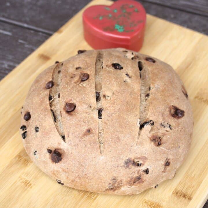 A round loaf of pumpernickel raisin bread on a cutting board with a red heart container in the background.