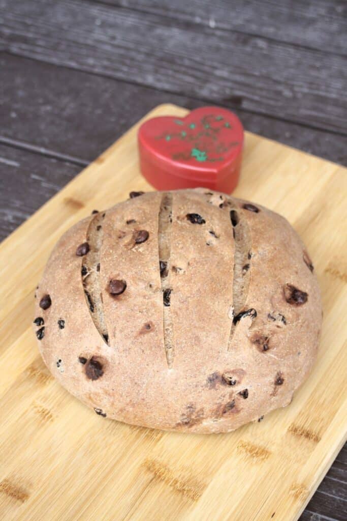 A round loaf of pumpernickel raisin bread on a wood cutting board with a red heart shaped container sitting behind it.