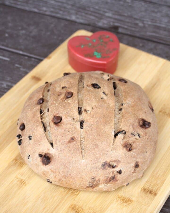 A round loaf of pumpernickel raisin bread on a wood cutting board with a red heart shaped container sitting behind it.