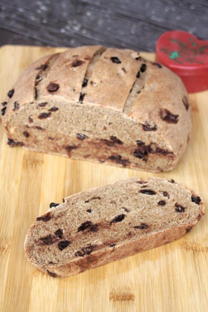 A slice of pumpernickel raisin bread on a wooden cutting board with remaining loaf and red heart shaped container in the background.