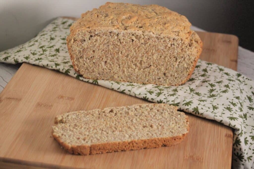 A slice of irish oatmeal soda bread sitting on a cutting board with remaining loaf sitting on a green and white linen in the background.