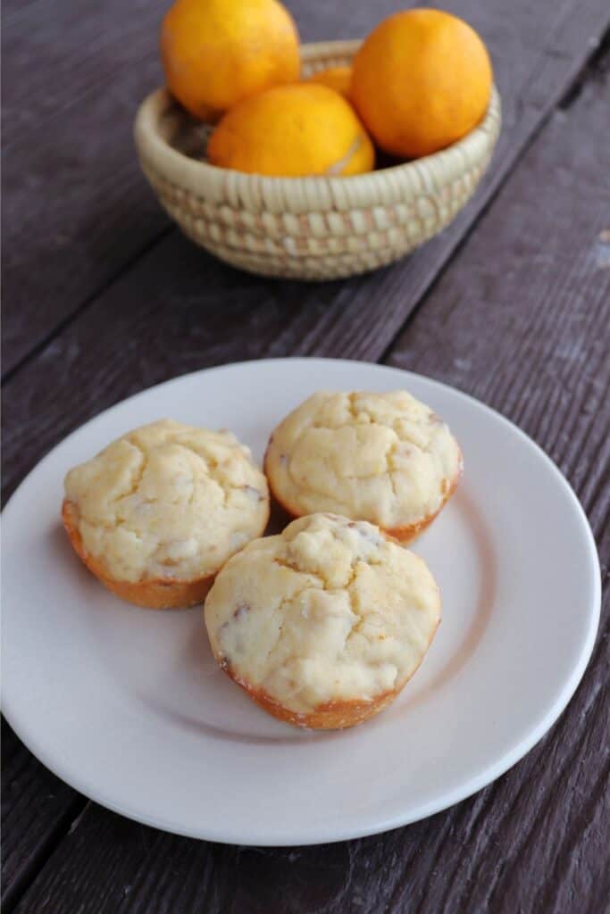 3 lemon nut muffins on a white plate with a basket of fresh meyer lemons behind it.