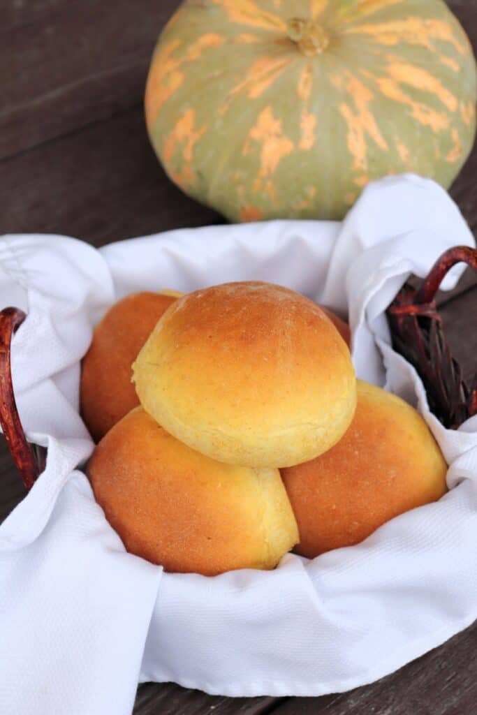 A white napkin lined basket full of pumpkin dinner rolls sitting in front of a green and orange striped pumpkin.