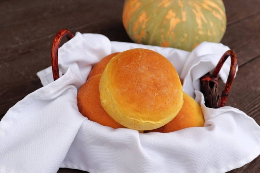 A white napkin lined basket full of pumpkin dinner rolls sitting in front of a green and orange striped pumpkin.