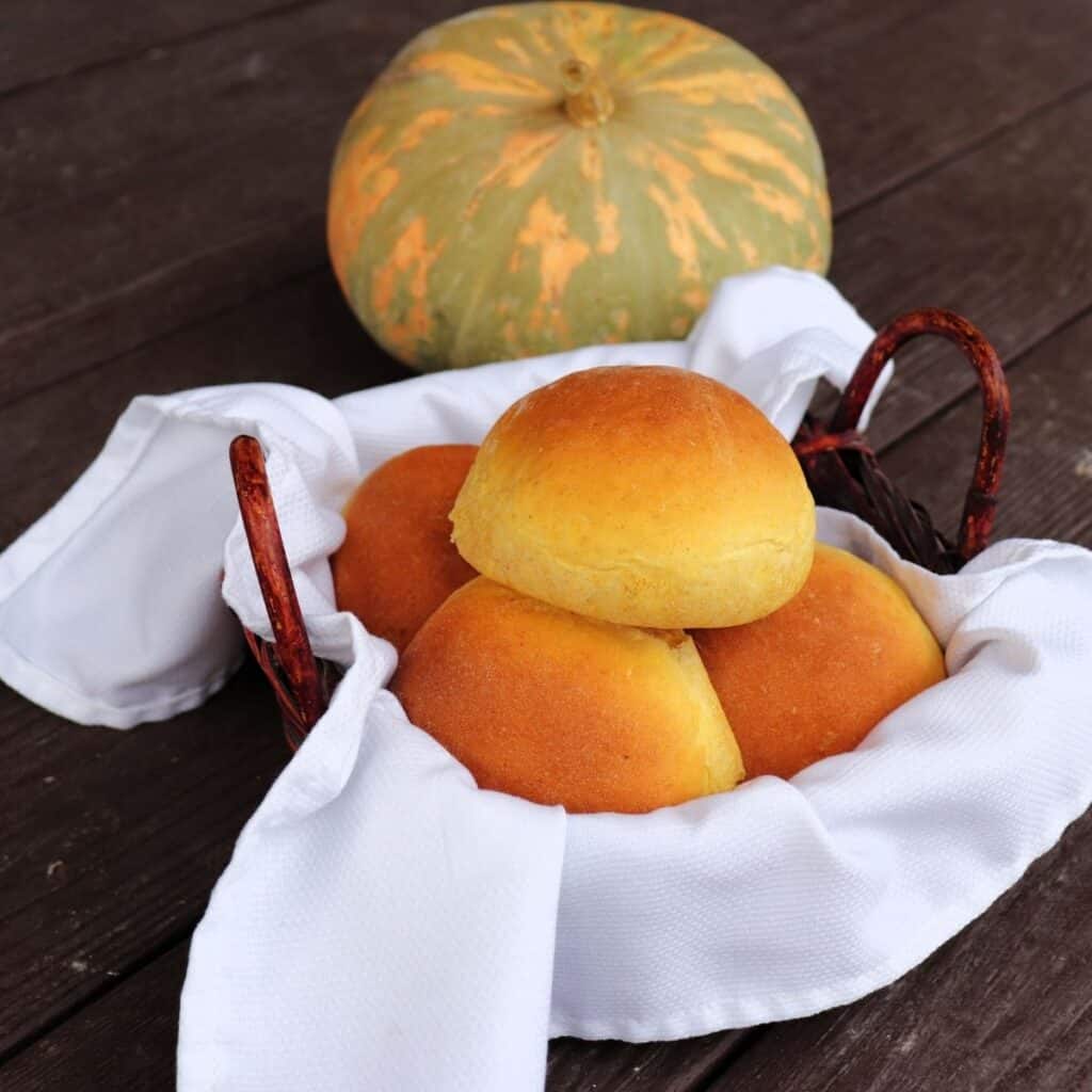 A white napkin lined basket full of pumpkin dinner rolls sitting in front of a green and orange striped pumpkin.