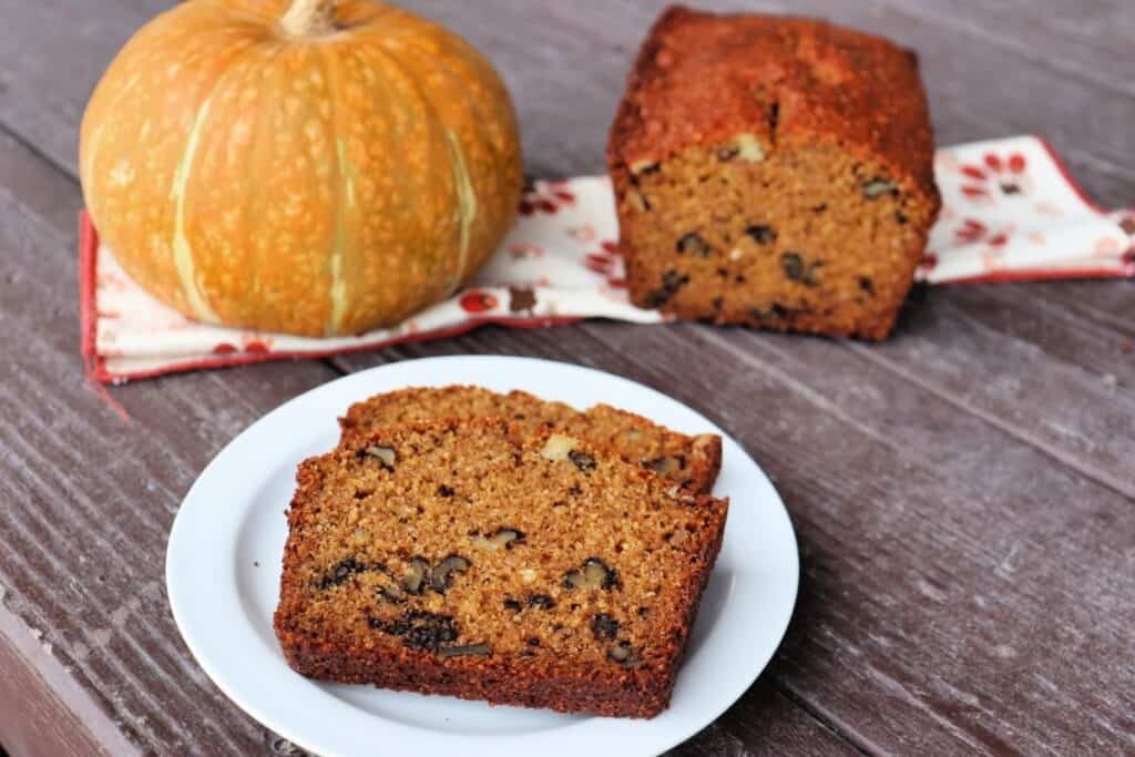 2 slices of whole wheat pumpkin bread on a white plate with rest of loaf and a pumpkin sitting on a napkin in the background.