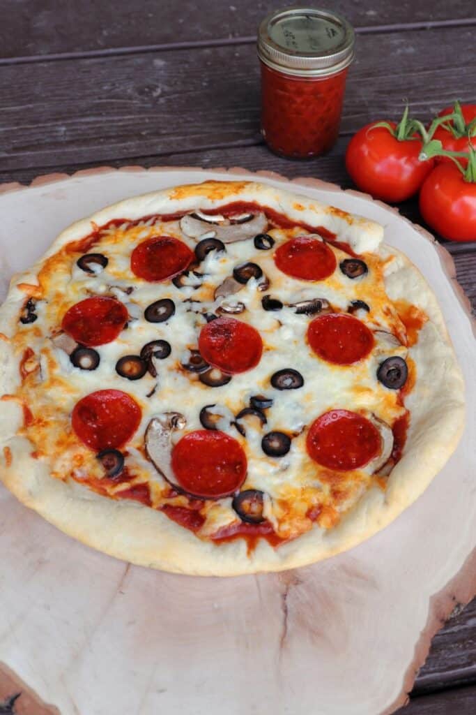 A pizza on a round wooden board with a jar of pizza sauce and fresh tomatoes in the background.
