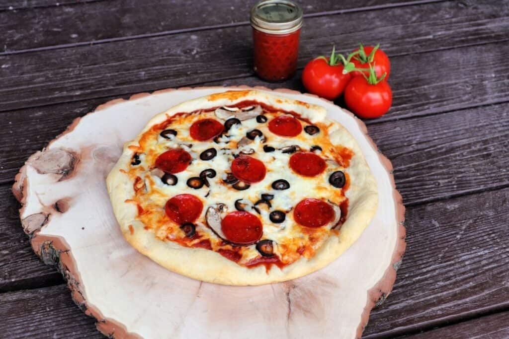 A pizza on a round wooden board with a jar of pizza sauce and fresh tomatoes in the background.