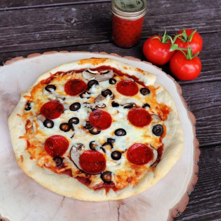 A pizza on a round wooden board with a jar of pizza sauce and fresh tomatoes in the background.