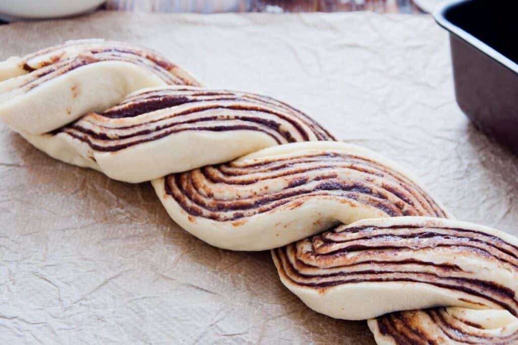 Bread dough halves exposing layers of jam twisted into 1 raw loaf.