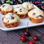 5 sour cherry chocolate chunk muffins on a square white plate with fresh cherries in front and a black cloth covered in a red cherry pattern behind.