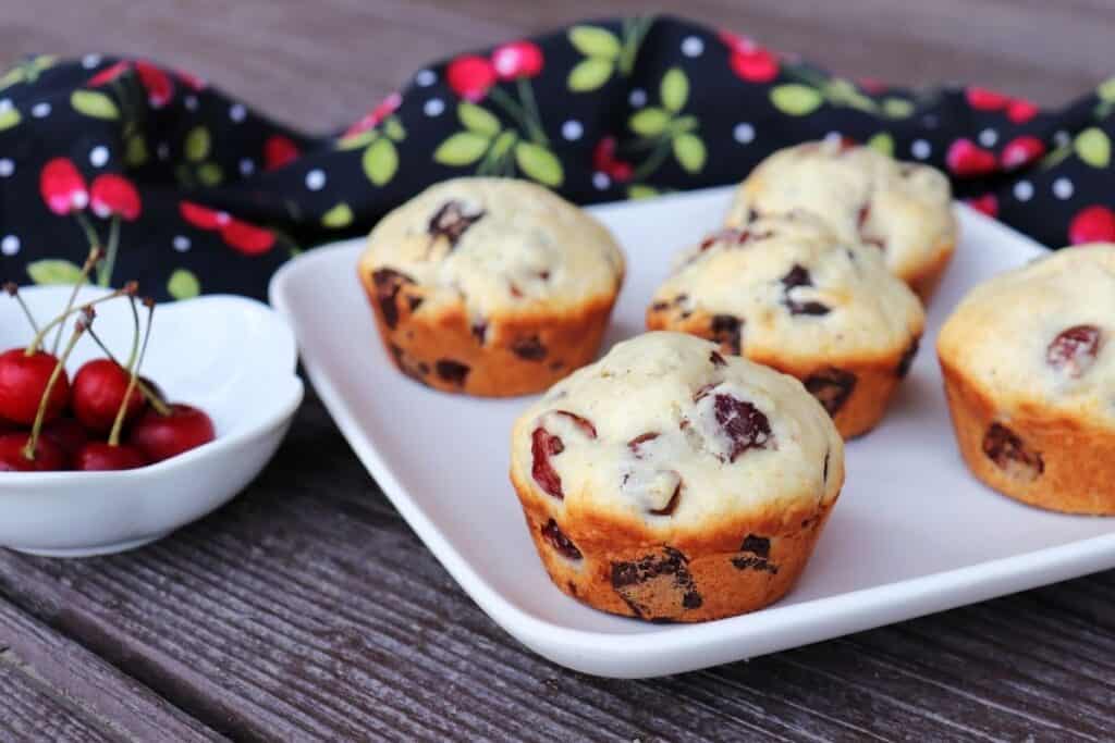 Sour cherry chocolate chunk muffins on a white plate with a bowl of fresh cherries sitting to the right. 