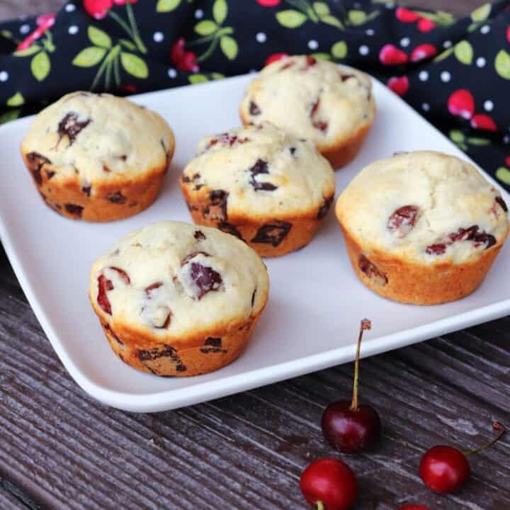 5 sour cherry chocolate chunk muffins on a square white plate with fresh cherries in front and a black cloth covered in a red cherry pattern behind.