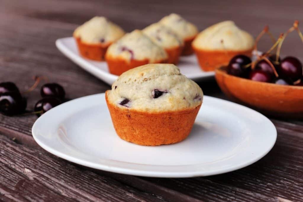 A muffins sitting on a white plate with a platter of more muffins in the background and a wooden bowl full of sweet cherries beside it.