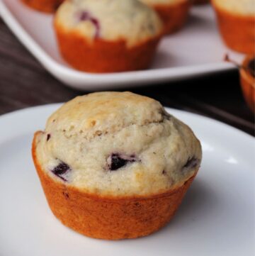 A cherry muffin on a white plate with platter of more muffins in the background.
