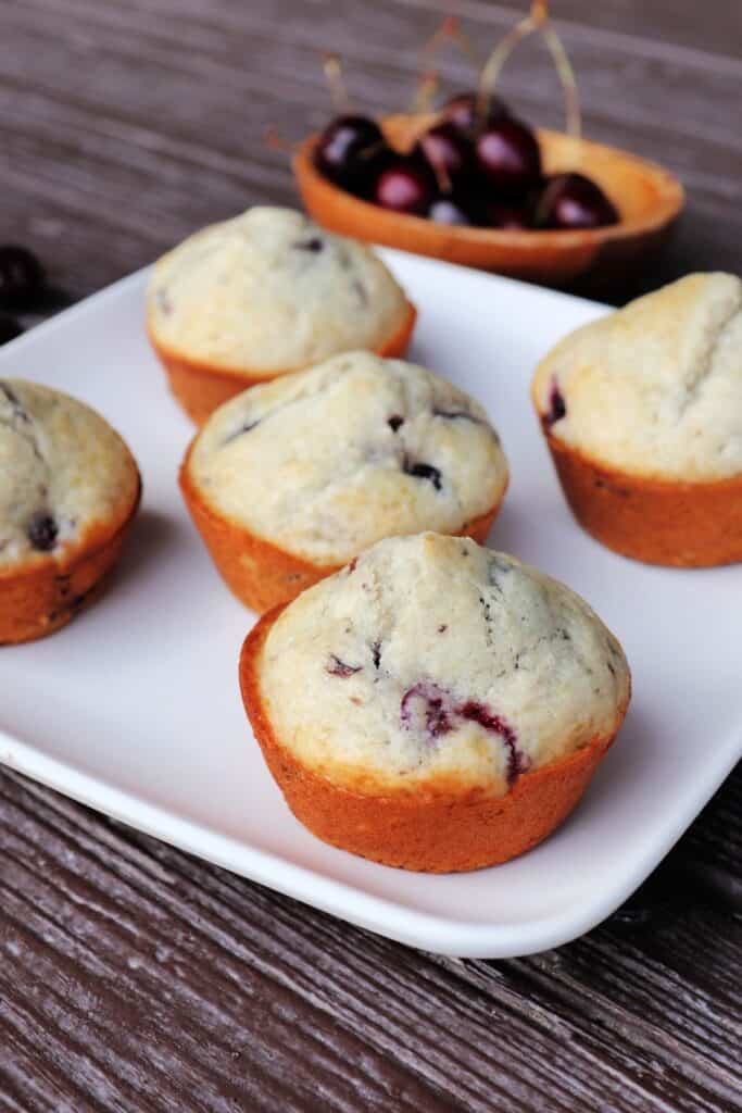 A square white plate with five muffins sitting on it, a wooden bowl of fresh cherries in the background.