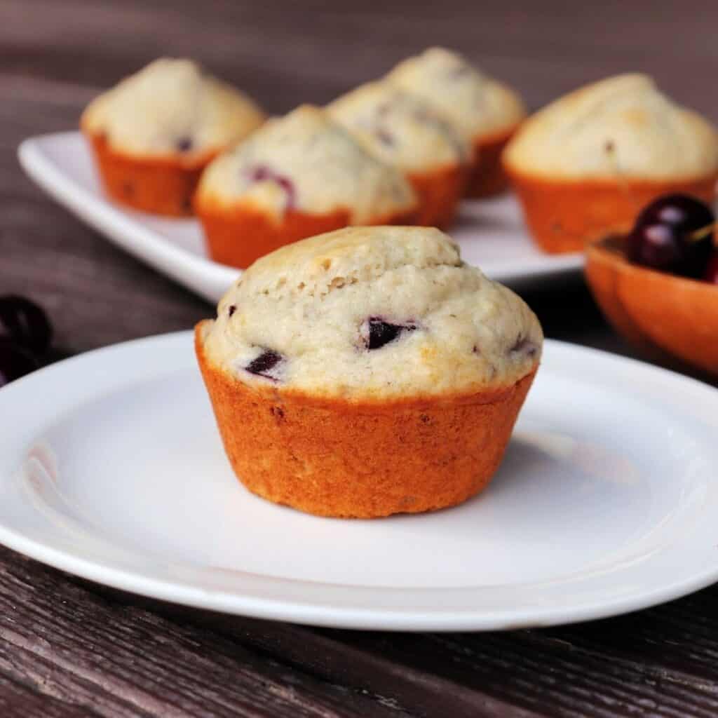 A cherry muffin on a white plate with platter of more muffins in the background.