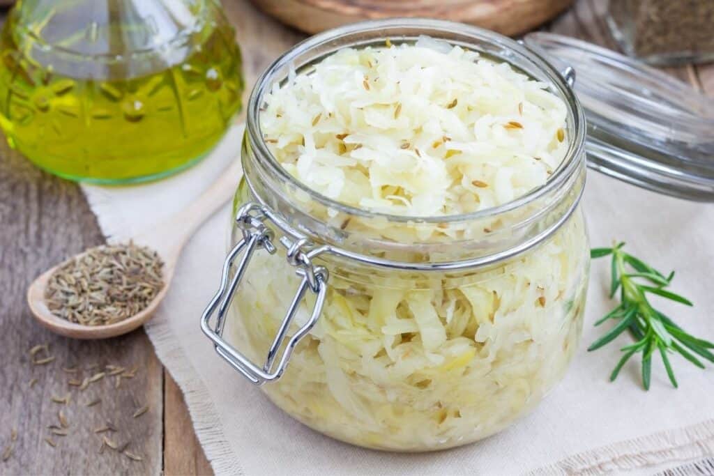 An open jar of sauerkraut sitting on a white napkin with a spoonful of seeds sitting next to it. 