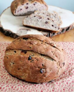 A round loaf of cranberry wild bread sitting on a brown and read cloth with another loaf of bread and slices sitting on a wooden board behind it.