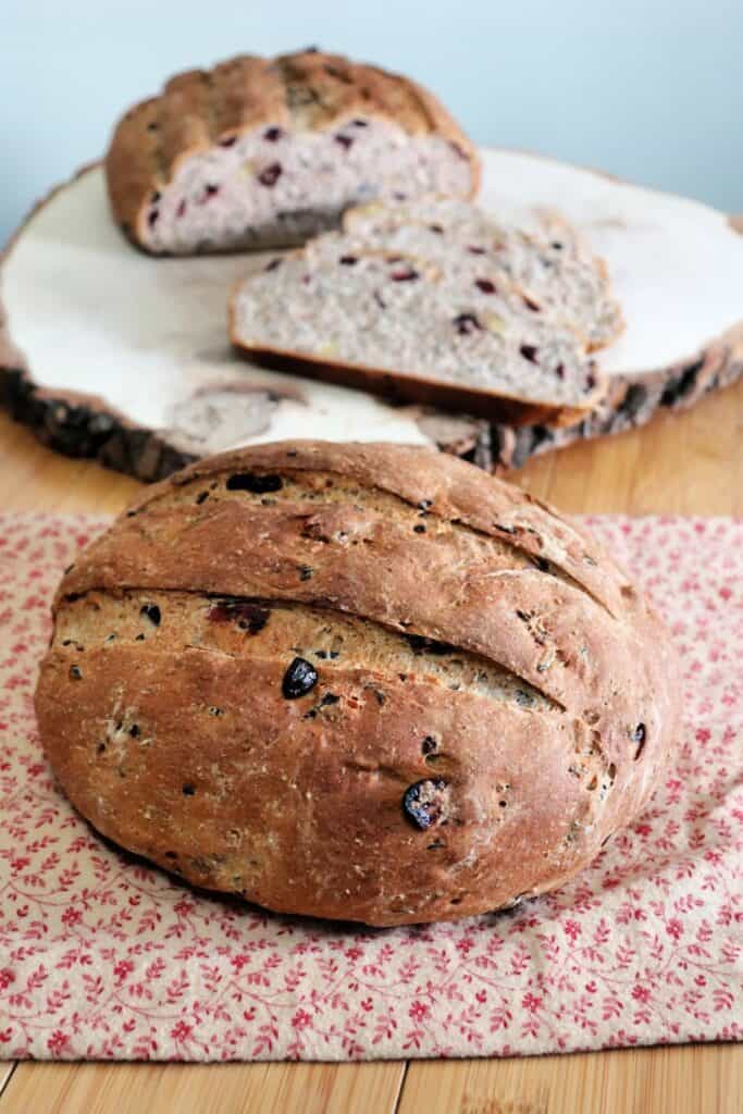 A round loaf of cranberry wild bread sitting on a brown and read cloth with another loaf of bread and slices sitting on a wooden board behind it.
