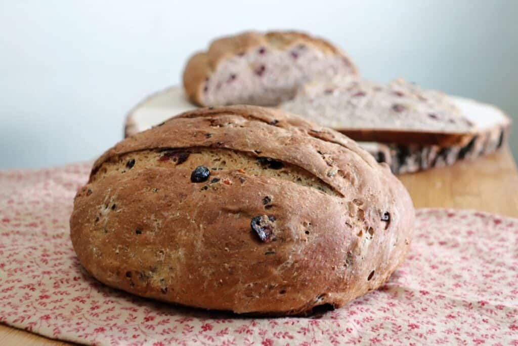 A round loaf of cranberry wild rice bread sitting on a brown and read cloth with another loaf of bread and slices sitting on a wooden board behind it. 