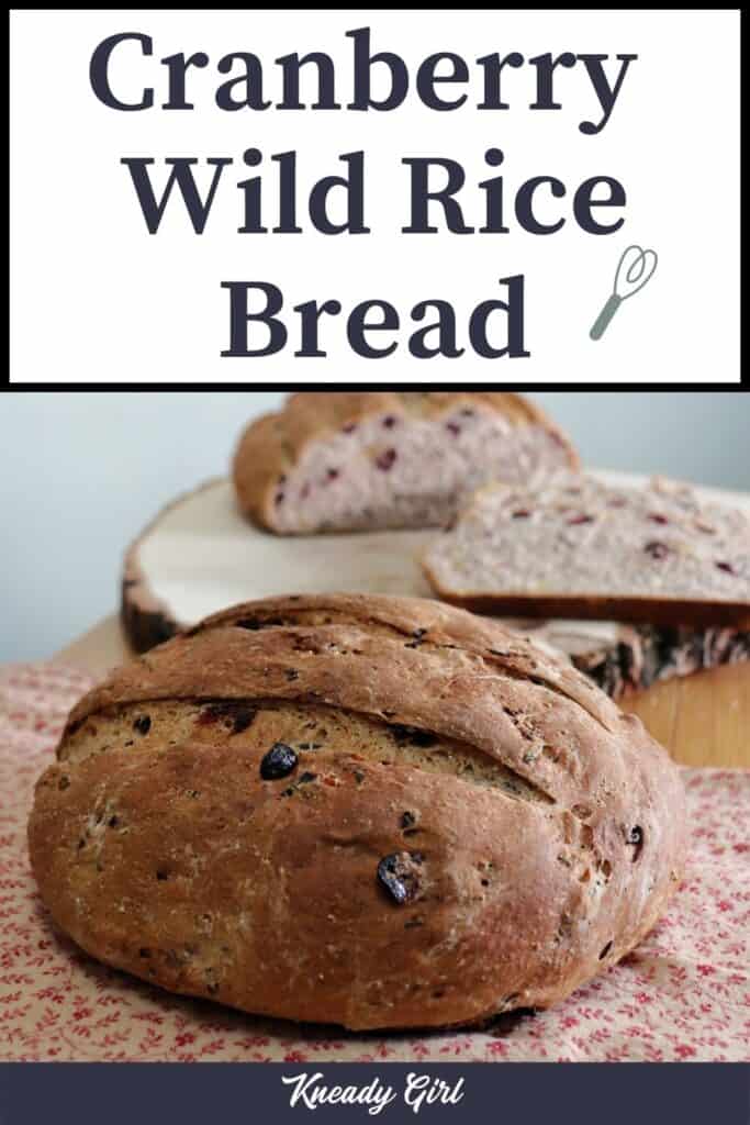 A round loaf of bread sitting on a brown and read cloth with another loaf of bread and slices sitting on a wooden board behind it. Text overlay reads: Cranberry wild rice bread.