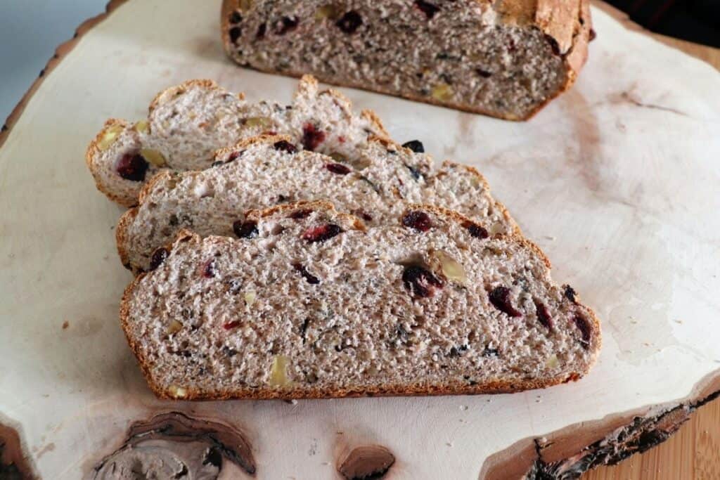 3 slices of cranberry wild rice bread on a wooden board with remaining loaf sitting behind it. 