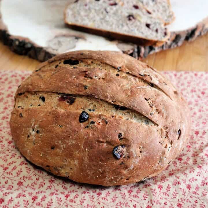 A round loaf of cranberry wild ricebread sitting on a brown and read cloth with another loaf of bread and slices sitting on a wooden board behind it.