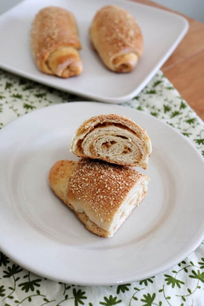A senorita bread roll sliced in half and facing up towards the camera sitting on a white plate. A plate with 2 whole rolls sits in the background. 