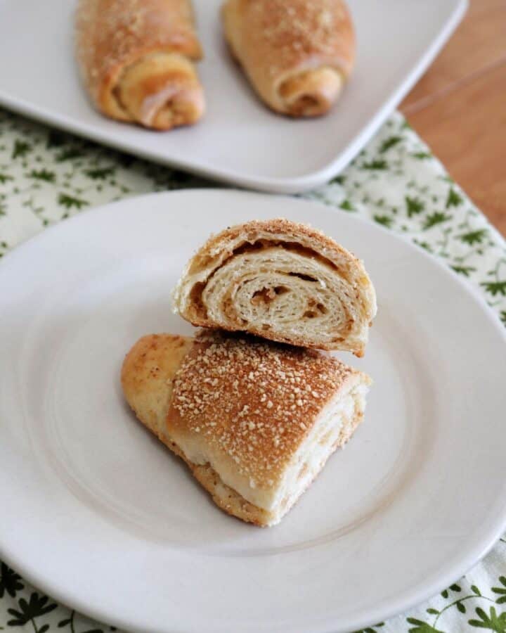 A senorita bread roll sliced in half and facing up towards the camera sitting on a white plate. A plate with 2 whole rolls sits in the background.