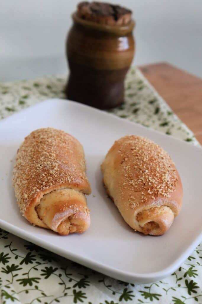 Two Señorita Bread rolls as seen from above sitting on a square white plate on top of a green and white cloth with a brown clay jar in the background.