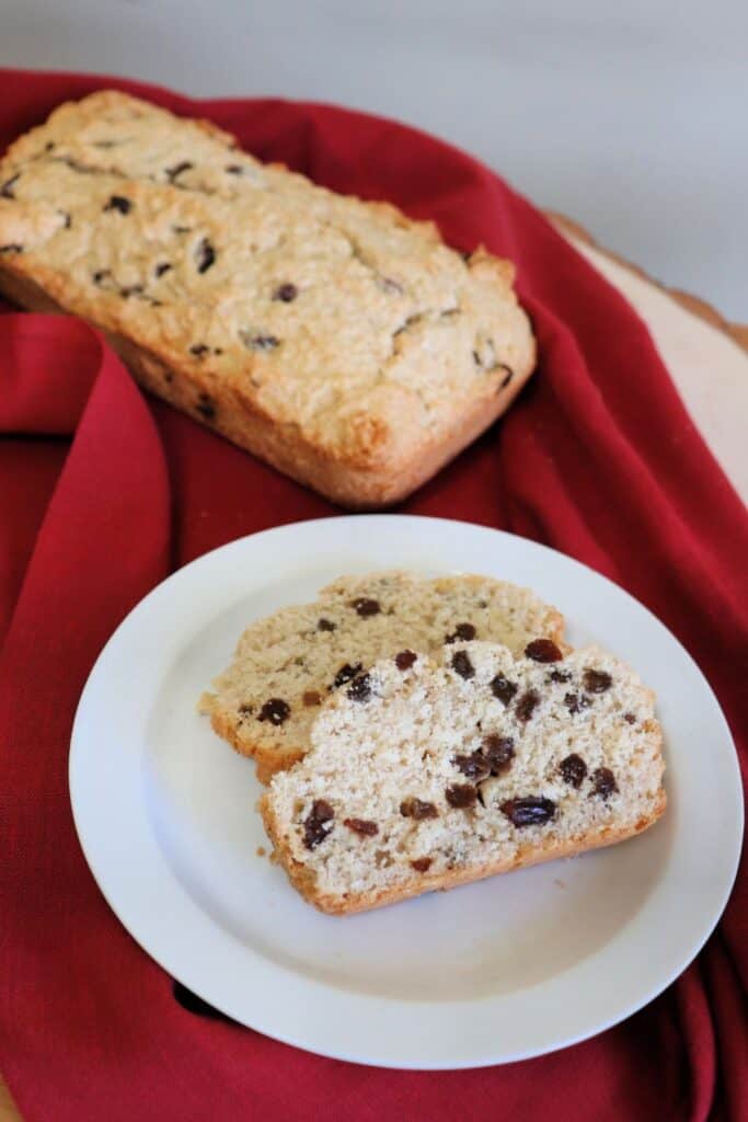 2 slices of coconut bread sitting on a white plate on top of a red cloth with a a whole loaf behind it.