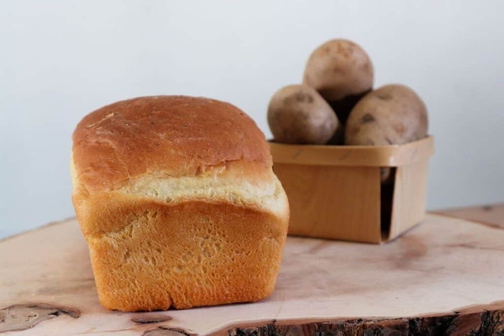 A loaf of potato bread on a board with a small basket overflowing with potatoes in the background. 