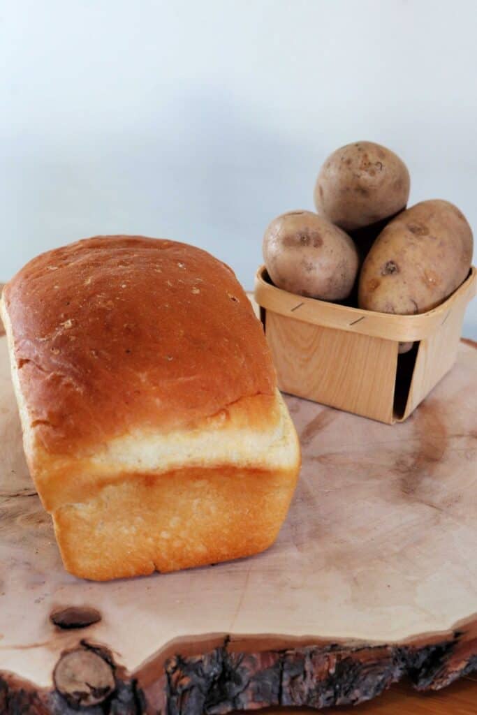 A loaf of potato bread on a board with a small basket overflowing with potatoes in the background. 