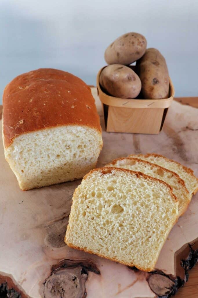 Slices of white bread on a wood board with remaining loaf to the left and a basket of potatoes in the background. 