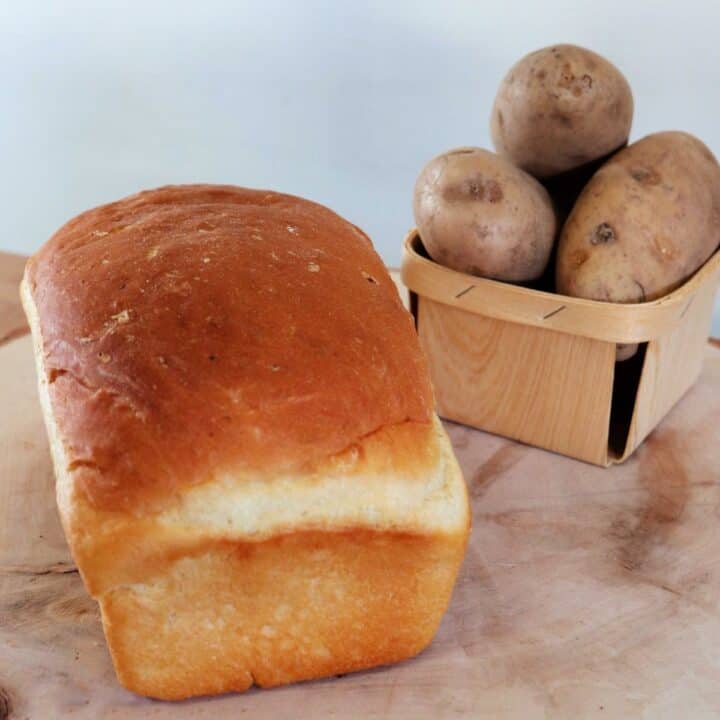 A loaf of potato bread on a board with a small basket overflowing with potatoes in the background.