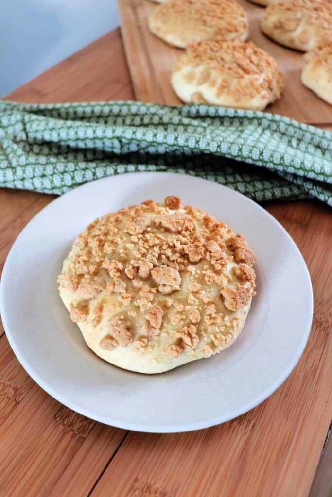 A streusel topped white bread roll sitting on a white plate with a green cloth behind it and more bread on a tray in the background.