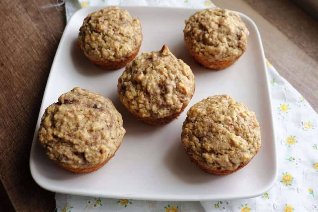 Banana bran muffins sitting on a white plate that is sitting on a white and yellow floral cloth as seen from above.
