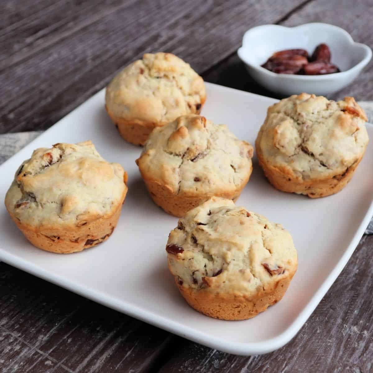 5 date muffins on a white plate with a bowl of dates in the background.