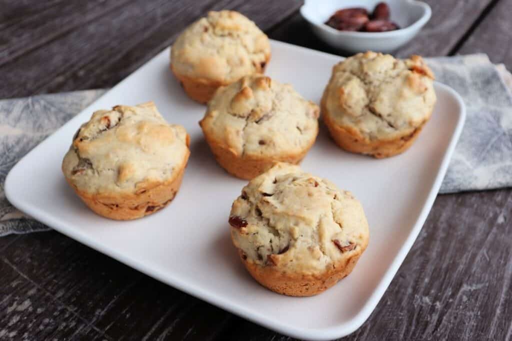 Date nut muffins on a square white plate sitting at an angle on a wooden table. with a bowl of dates in the background.