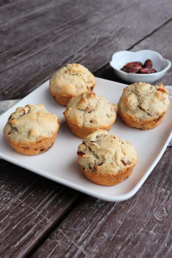 Date nut muffins on a square white plate sitting at an angle on a wooden table. with a bowl of dates in the background.