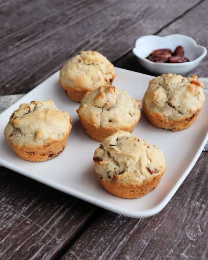 Date nut muffins on a square white plate sitting at an angle on a wooden table. with a bowl of dates in the background.
