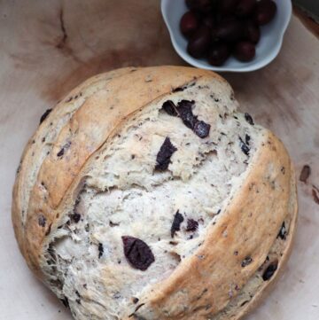A loaf of kalamata olive bread as seen from above sitting on a wood board with a bowl of olives sitting next to it.